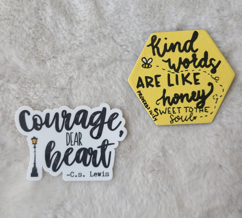 May include: Two yellow and black motivational stickers. The first sticker is a hexagon with the text "Kind words are like honey sweet to the soul" and a small bee illustration. The second sticker is a white background with black text that reads "Courage, dear heart - C.S. Lewis" and a black silhouette of a street lamp.