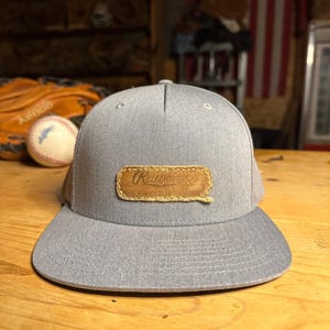 May include: A heather grey baseball cap with a flat brim and a rectangular tan leather patch stitched onto the front. The patch features the embossed text Rawlings and The Finest in the Field, with visible stitching around the leather edges.