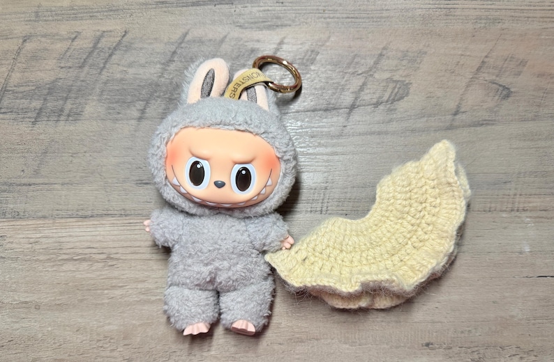 May include: A gray plush bunny figure with a mischievous expression, wearing a matching hooded outfit. A separate, light yellow crocheted item rests beside it. The figure has a gold-colored keychain attached.
