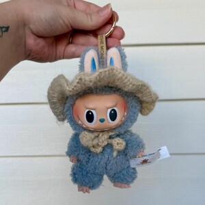 May include: A blue plush toy keychain with a bunny hat and a beige knitted hat. The toy has a round face with large eyes and a small tag. The keychain ring is gold.