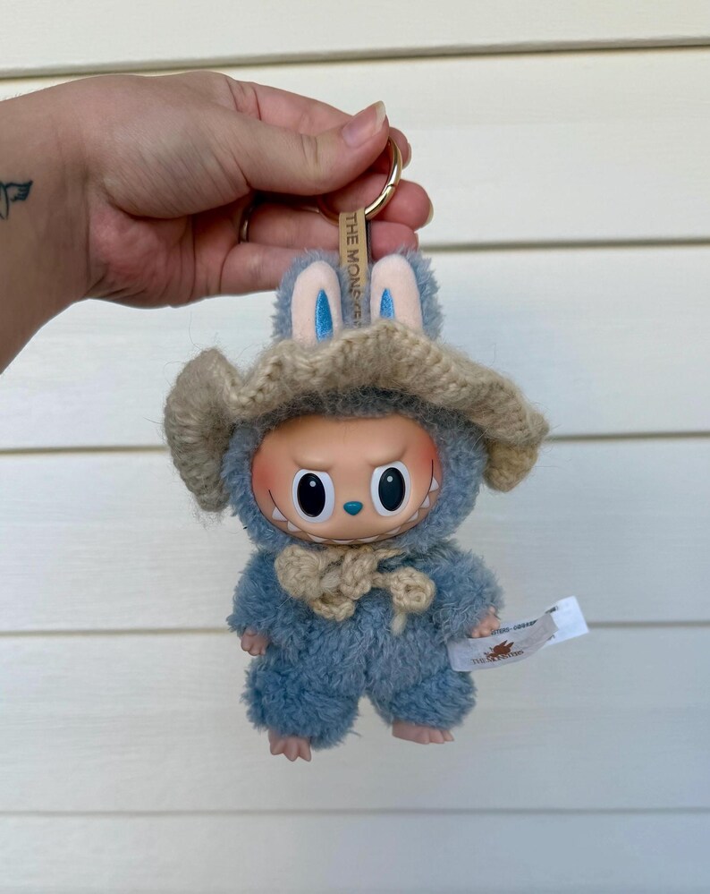 May include: A blue plush toy keychain with a bunny hat and a beige knitted hat. The toy has a round face with large eyes and a small tag. The keychain ring is gold.
