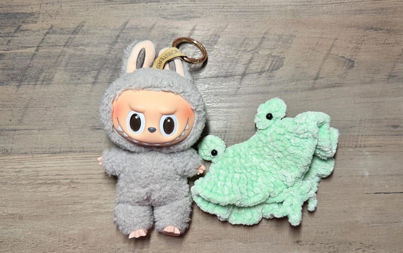 K&ouml;nnte beinhalten: Eine Pl&uuml;schpuppe mit grauen Hasenohren und einem schelmischen Gesichtsausdruck sowie ein hellgr&uuml;ner geh&auml;kelter Frosch. Die Puppe hat einen Schl&uuml;sselanh&auml;nger. Der Frosch ist hellgr&uuml;n.