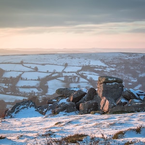 Puede incluir: Una vista panorámica de un paisaje cubierto de nieve con afloramientos rocosos en primer plano. Las colinas distantes están cubiertas de nieve y el cielo es de un suave rosa y azul.