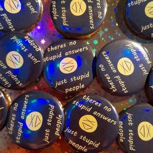 May include: A collection of dark blue pinback buttons with the text "there's no stupid answers just stupid people" in white. Each button has a yellow circle with the letters "XD" inside. The buttons are arranged on a reflective surface.