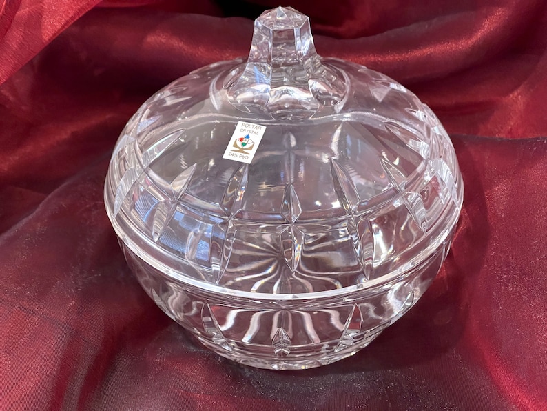 May include: A clear glass candy dish with a lid. The dish has a cut glass design and is decorated with a label that reads "Poltar Crystal 24% PbO".