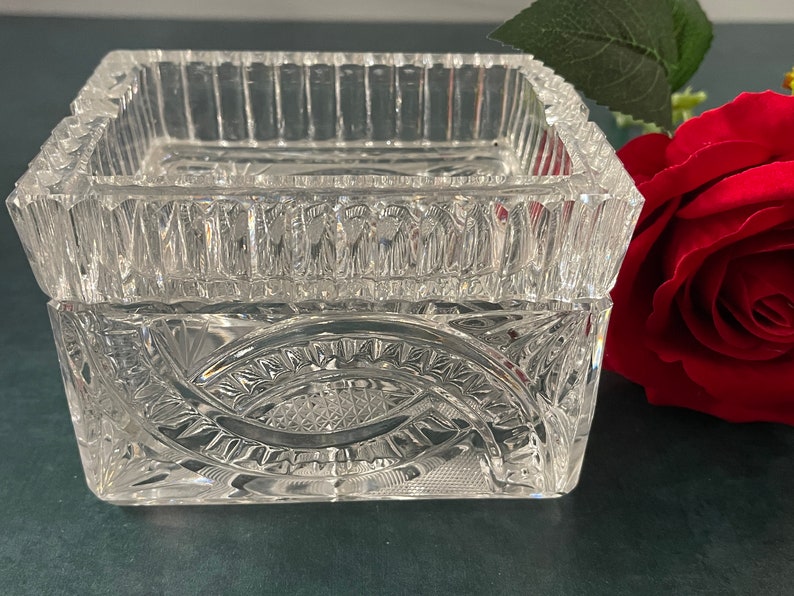 Op de afbeelding: Een vierkante doos van helder glas met ingewikkelde geslepen glasontwerpen. De doos heeft een verhoogde rand en een decoratief patroon op de bodem.
