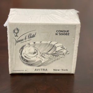 May include: A white box with a clear glass shell-shaped dish inside. The box has black text that reads "Yannes le Chatel Collection France" and "CONQUE N°55082". The bottom of the box reads "DISTRIBUTED BY AVITRA New-York".