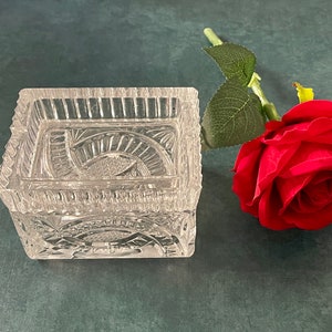 May include: A clear glass decorative box with intricate cut-out designs. The box is sitting on a dark blue surface next to a single red rose.