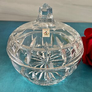 May include: A clear glass candy dish with a lid and a diamond-shaped knob. The dish has a cut glass design and is sitting on a blue surface. The label on the dish reads "Poltar Crystal 24% PbO."