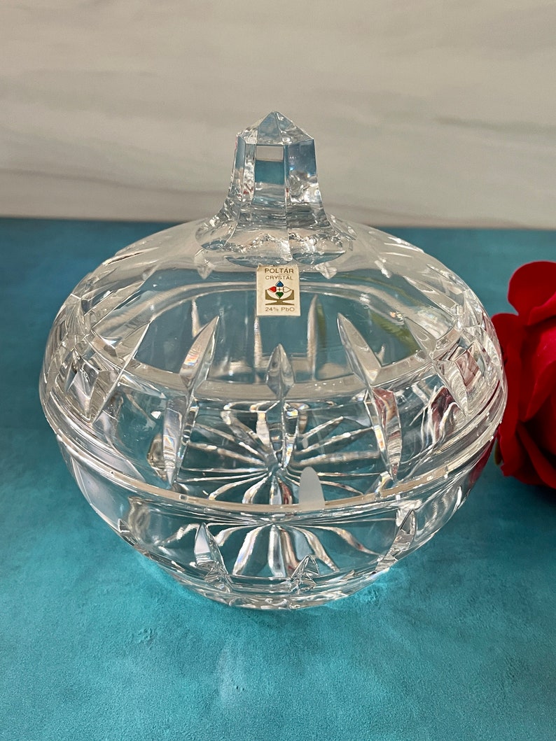 May include: A clear glass candy dish with a lid and a diamond-shaped knob. The dish has a cut glass design and is sitting on a blue surface. The label on the dish reads "Poltar Crystal 24% PbO."