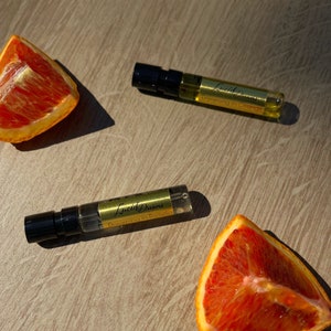 May include: Two small glass bottles of perfume with black caps and gold labels. The labels read "Lucid Dreams" and "Dreaming in Technicolor". The bottles are on a wooden surface with two slices of red orange.