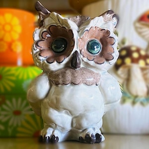 May include: A ceramic owl figurine with a white body, brown and tan accents, and large, round, teal-colored eyes. The owl has a detailed, textured appearance and is standing upright. Other ceramic items are visible in the background.