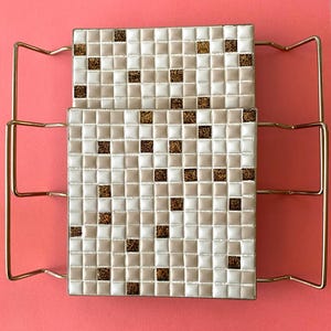 May include: A two-tiered, gold-toned metal rack with square ceramic tiles. The tiles are a mosaic of white, beige, and brown squares. The rack is set against a pink background, creating a vintage aesthetic.