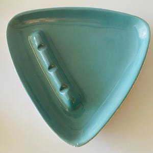May include: A turquoise ceramic ashtray in a triangular shape. The ashtray has a raised section with three grooves for resting cigarettes. The edges have a slightly distressed, off-white finish.