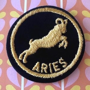 May include: A black circular embroidered patch featuring a gold ram leaping within a gold stitched border. The word "ARIES" is embroidered in gold below the ram. The patch is on a patterned background.