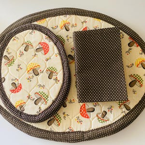 May include: A large oval quilted placemat and a smaller oval quilted hot pad, both featuring a cream background with a pattern of colorful mushrooms and small flowers, finished with a dark brown polka dot fabric border and a matching folded napkin.