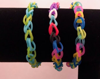 May include: Three colorful rubber band bracelets on a black display stand. The bracelets are made with different colored rubber bands in a woven pattern. The bracelets are green and blue, pink and blue, and yellow and blue.