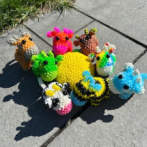 May include: A collection of colorful, handmade loom-band animal figures. The assortment includes a yellow sphere, a blue fish, a bee, a cow, and several other creatures in shades of orange, green, and pink. The figures are arranged on a gray surface.