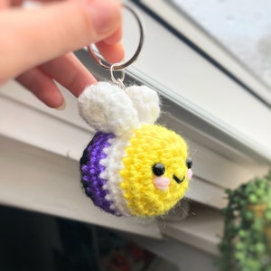 May include: A handmade crocheted bee keyring in yellow, white, purple, and black, with a silver ring. The bee has small black eyes and pink cheeks. The wings are white and fluffy. A cute and colourful accessory.