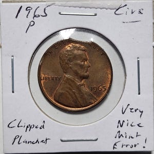 May include: A 1965 copper penny with a clipped planchet error, displayed in a protective holder. The coin features the profile of Abraham Lincoln and the text "Liberty" and "In God We Trust". The holder is labeled "Very Nice Mint Error!"