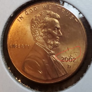 May include: A copper-colored US penny with the image of Abraham Lincoln on the front. The coin has the words "IN GOD WE TRUST" and "LIBERTY" inscribed on it. The year "2002" and the letter "D" are visible on the bottom of the coin.