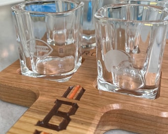May include: Wooden shot glass holder with four shot glasses. The holder is engraved with the word "Browns" and has orange and brown stripes. The shot glasses are clear and have a football helmet design.