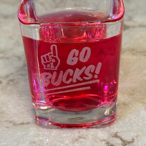 May include: A clear shot glass filled with a red liquid. The glass has the words "GO BUCKS!" etched in white, along with a hand symbol. The glass is sitting on a speckled surface.