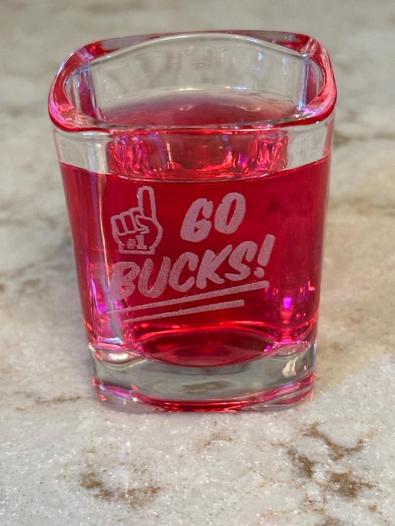 May include: A clear shot glass filled with a red liquid. The glass has the words "GO BUCKS!" etched in white, along with a hand symbol. The glass is sitting on a speckled surface.