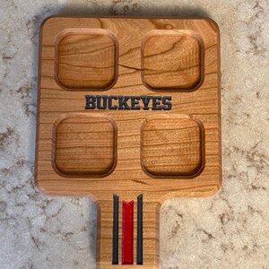 May include: Wooden serving tray with four square indentations and a handle. The word "BUCKEYES" is carved into the wood. The handle features black and red stripes. The tray is made of light-colored wood.