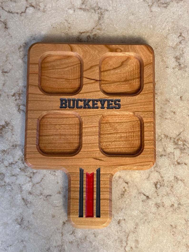 May include: Wooden serving tray with four square indentations and a handle. The word "BUCKEYES" is carved into the wood. The handle features black and red stripes. The tray is made of light-colored wood.