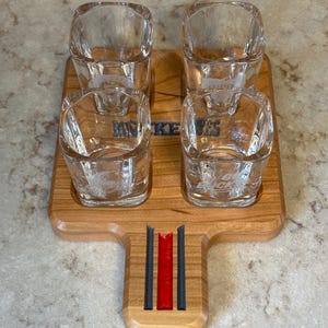 May include: A wooden shot glass serving tray with four square shot glasses. The tray has a handle with red and black stripes. The shot glasses and tray are etched with the word "Buckeyes".