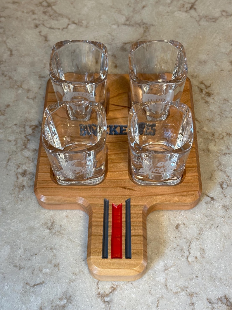 May include: A wooden shot glass serving tray with four square shot glasses. The tray has a handle with red and black stripes. The shot glasses and tray are etched with the word "Buckeyes".
