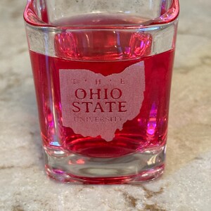 May include: A clear, square shot glass filled with a vibrant red liquid. The glass features an etched design of the state of Ohio with the words "OHIO STATE UNIVERSITY". The shot glass is sitting on a light-colored surface.