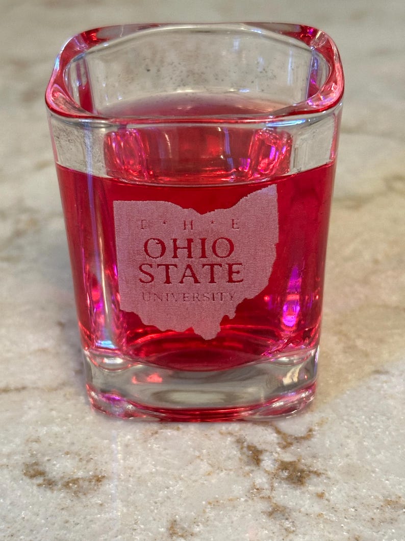 May include: A clear, square shot glass filled with a vibrant red liquid. The glass features an etched design of the state of Ohio with the words "OHIO STATE UNIVERSITY". The shot glass is sitting on a light-colored surface.
