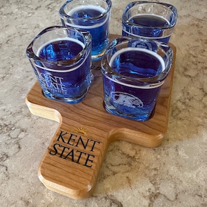 May include: A wooden shot glass holder with four shot glasses filled with blue liquid. The holder is engraved with the words "Kent State".