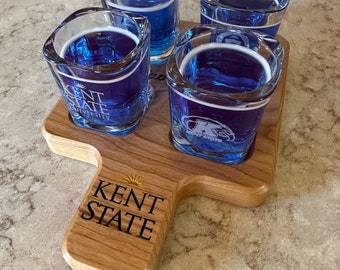 May include: A wooden shot glass holder with four shot glasses filled with blue liquid. The holder is engraved with the words "Kent State".