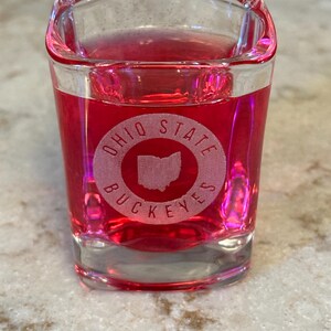 May include: A clear, square shot glass filled with a red liquid. The glass features the words "OHIO STATE BUCKEYES" in a circular design, with the outline of the state of Ohio in the center. The glass is sitting on a light-colored surface.