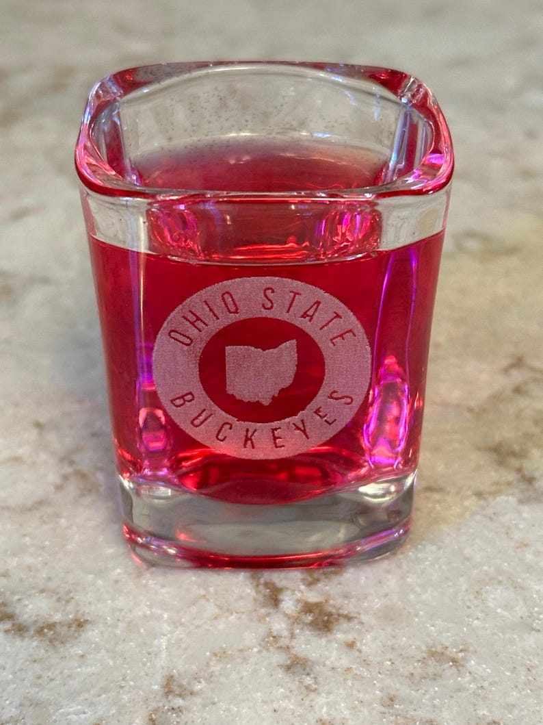 May include: A clear, square shot glass filled with a red liquid. The glass features the words "OHIO STATE BUCKEYES" in a circular design, with the outline of the state of Ohio in the center. The glass is sitting on a light-colored surface.