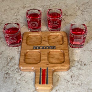 May include: A wooden shot glass serving tray with four square cutouts, holding four shot glasses filled with red liquid. The tray has the word "BUCKEYES" engraved on it. The shot glasses have the words "GO BUCKS!" and "OHIO STATE" printed on them.