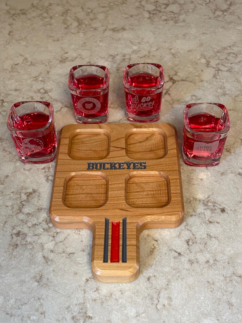 May include: A wooden shot glass serving tray with four square cutouts, holding four shot glasses filled with red liquid. The tray has the word "BUCKEYES" engraved on it. The shot glasses have the words "GO BUCKS!" and "OHIO STATE" printed on them.