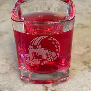 May include: A clear shot glass filled with a red liquid. The glass features an etched design of a football helmet. The shot glass is square-shaped with a slightly curved top edge. The glass is sitting on a light-colored surface.