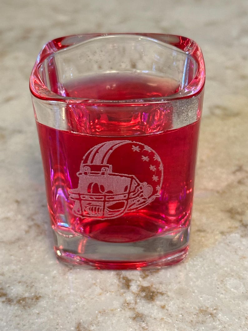 May include: A clear shot glass filled with a red liquid. The glass features an etched design of a football helmet. The shot glass is square-shaped with a slightly curved top edge. The glass is sitting on a light-colored surface.