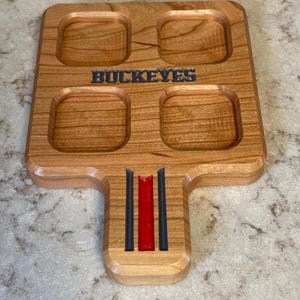 May include: Wooden serving tray with four square indentations and a handle. The word "BUCKEYES" is carved into the wood. The handle features red and black stripes. The tray is made of light brown wood.