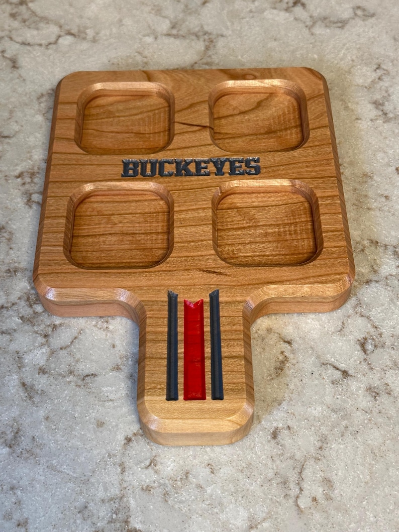 May include: Wooden serving tray with four square indentations and a handle. The word "BUCKEYES" is carved into the wood. The handle features red and black stripes. The tray is made of light brown wood.