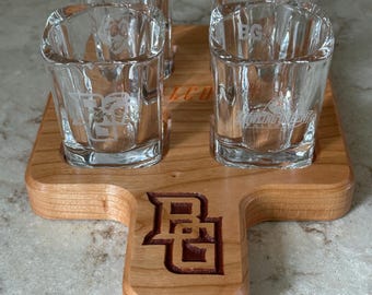 May include: A wooden shot glass serving tray with four clear square shot glasses. The tray has a handle and a logo etched into the wood. Each shot glass has a logo etched into the glass.