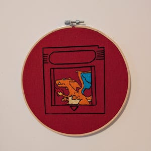 May include: Embroidered hoop art featuring a red background with a black outline of a video game cartridge. Inside the cartridge is an orange and blue dragon-like creature. The embroidery is set in a wooden hoop.