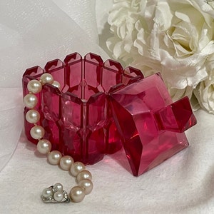 May include: A pink glass trinket box with a square shape and a faceted design. The box is open and a strand of white pearls is resting on the top of the box.