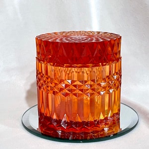 May include: An orange-colored, faceted glass jar with a matching lid. The jar sits on a small, round, mirrored surface. The glass has a diamond-like pattern, creating a textured appearance. The jar is cylindrical in shape.