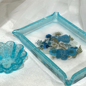 May include: A rectangular light blue resin tray with embedded dried blue flowers and green foliage, accompanied by a small, scalloped, light blue resin bowl featuring a textured, grid-like pattern on its exterior surface.