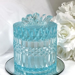 May include: A light blue, faceted glass jar with a decorative lid, resting on a small, round mirror. The jar has a geometric design and is surrounded by white flowers.